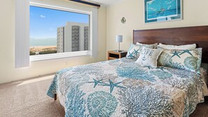 Une chambre avec une grande fenêtre, un lit recouvert d’une courtepointe aux motifs bleus et blancs évoquant l’océan, une table de chevet avec une lampe, et un tableau encadré représentant un bateau accroché au mur.