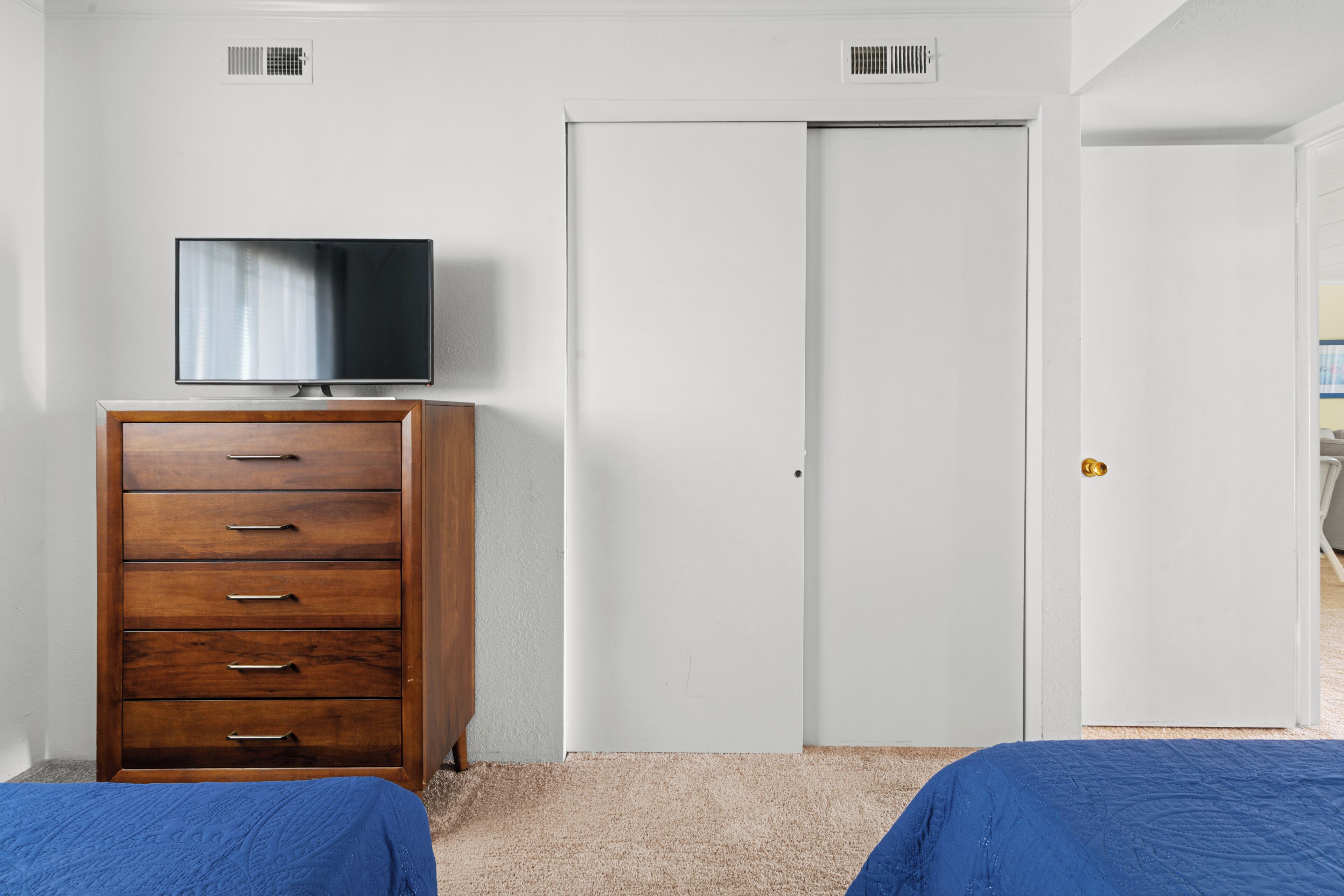 A bedroom with a wooden dresser, a television on top, a white door, and a bed with blue bedding.