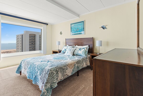 Une chambre à coucher comprenant un lit, des tables de chevet, un bureau, une fenêtre donnant sur un immeuble et la mer, ainsi qu’un tableau accroché au mur.