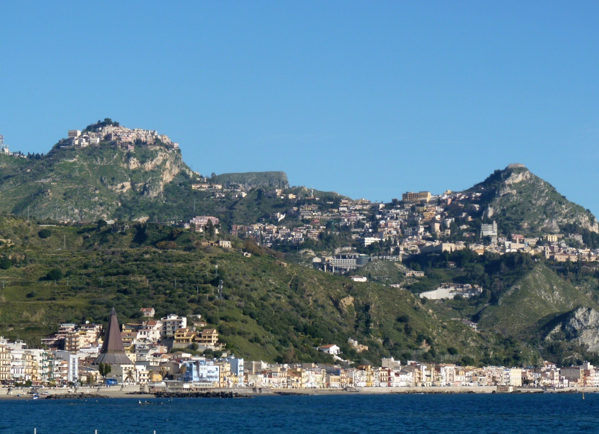A coastal town with a prominent hilltop fortress, dense urban buildings, and a waterfront area with visible structures.