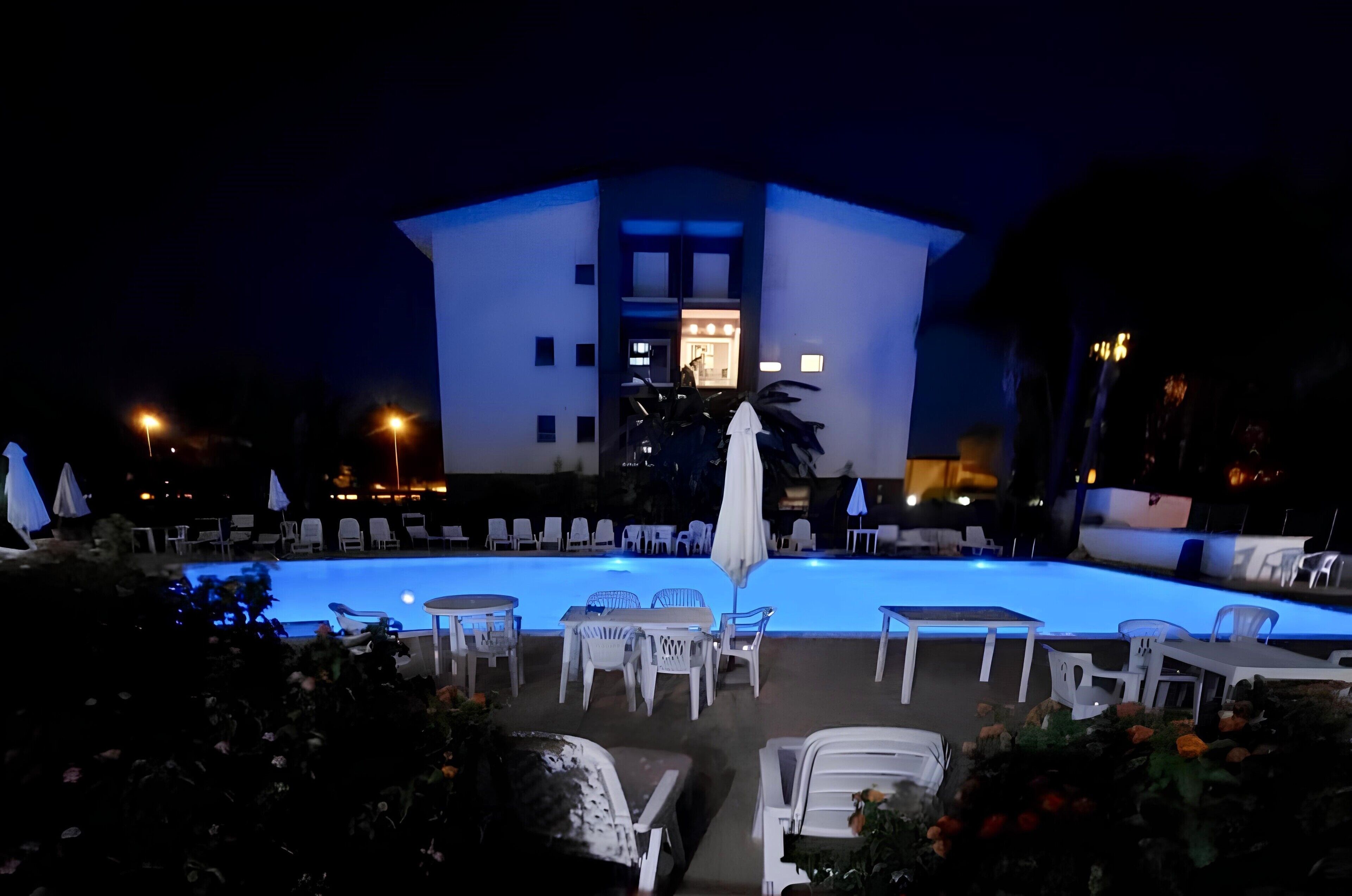 A nighttime view of a hotel with a swimming pool, outdoor seating, and a building with illuminated windows.