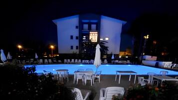 A nighttime view of a hotel with a swimming pool, outdoor seating, and a building with illuminated windows.