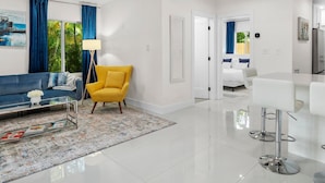 A modern living room with a blue sofa, a yellow armchair, a glass coffee table, and a patterned rug. - Uhost AI Stylish 1BR Bright Space w Patio (Fort Lauderdale)