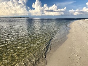 Een strand met helder ondiep water, een zandstrand en een verafgelegen landmassa onder een licht bewolkte lucht.