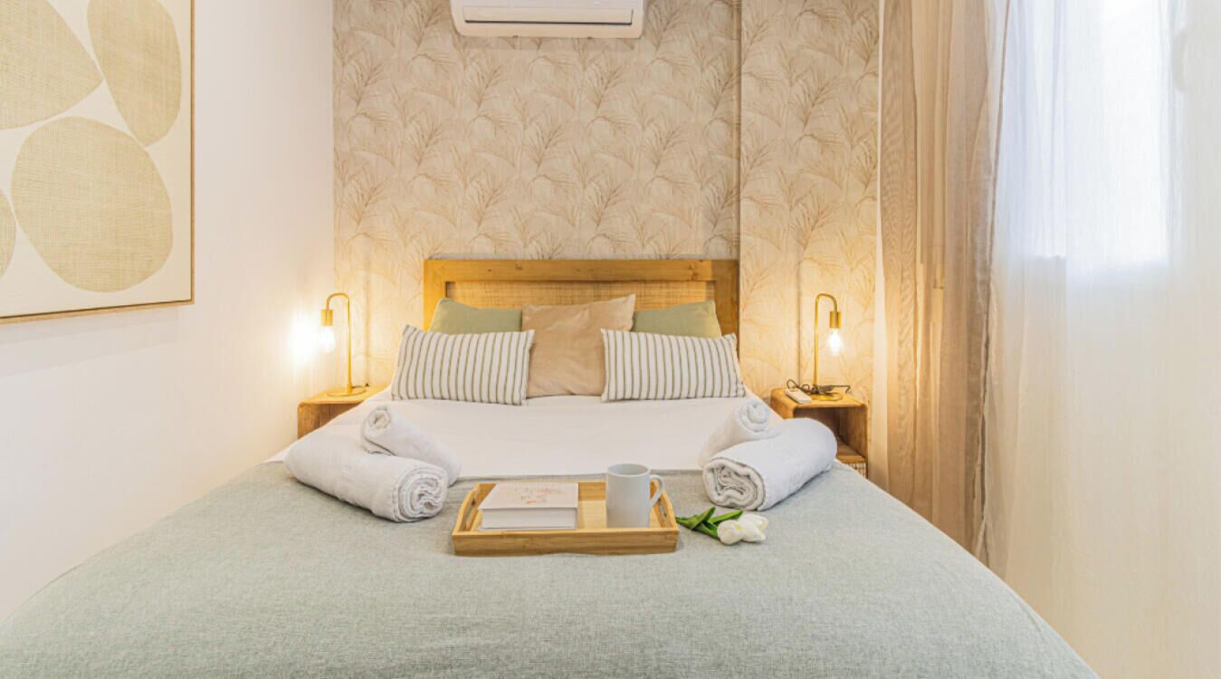 Una cama bien hecha con almohadas, una bandeja con un libro y una taza, y una flor en una habitación de hotel.