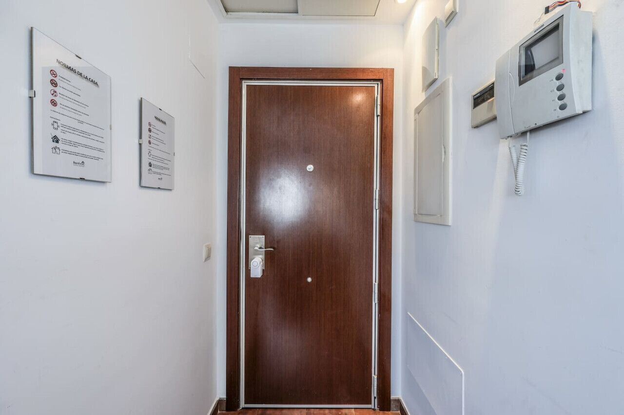 Entrada a una habitación de hotel con una puerta de madera, un portero automático en la pared y dos carteles informativos.