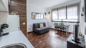Une cuisine moderne avec un plan de travail blanc, un four encastré et un évier. On y trouve un canapé gris avec un coussin coloré, une table à manger avec des chaises et une grande fenêtre avec des stores.