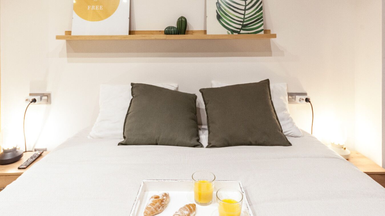 Een net opgemaakt bed met witte lakens, twee donkere kussens en een dienblad met croissants en sinaasappelsap.