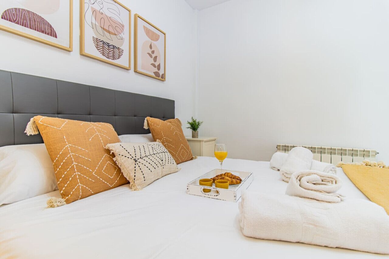 Una cama impecable con cabecera gris, ropa de cama blanca y almohadas decorativas. Hay una bandeja con cruasanes y un vaso de jugo de naranja sobre la cama. También se ven toallas blancas y una manta amarilla.