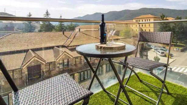 Un balcon avec une table et des chaises, d’où l’on peut admirer un paysage urbain avec des bâtiments et une montagne à l’horizon.