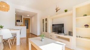 Un salon moderne doté d’un téléviseur à écran plat, d’un coin repas avec une table et des chaises blanches, et d’une cuisine visible au fond.