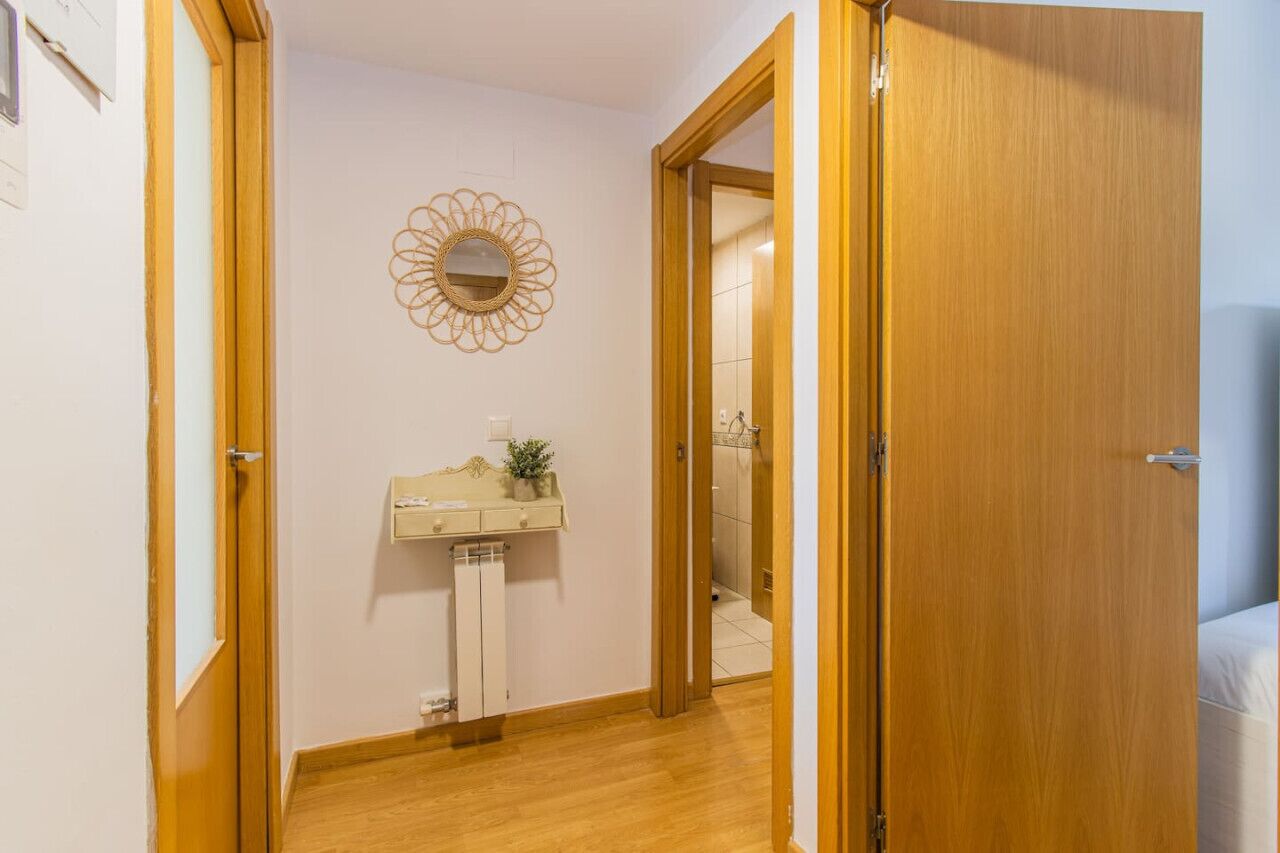 Un couloir étroit avec une porte en bois sur la droite, un miroir sur la gauche et un petit lavabo avec un porte-serviettes en dessous.