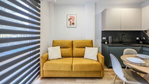 A modern living room with a sofa, a dining table with chairs, a kitchen area, and a large window with blinds. - Luxe Apartments Madrid Rio by Rentitup (Madrid)
