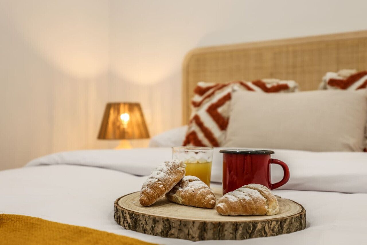 Un lit avec des oreillers, une lampe, des croissants, un verre de jus d’orange et une tasse rouge.