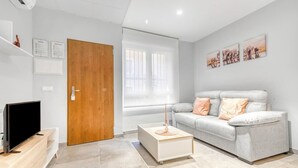 A modern living room with a grey sofa, a coffee table, a TV on a wooden stand, a wooden door, and a window with blinds.