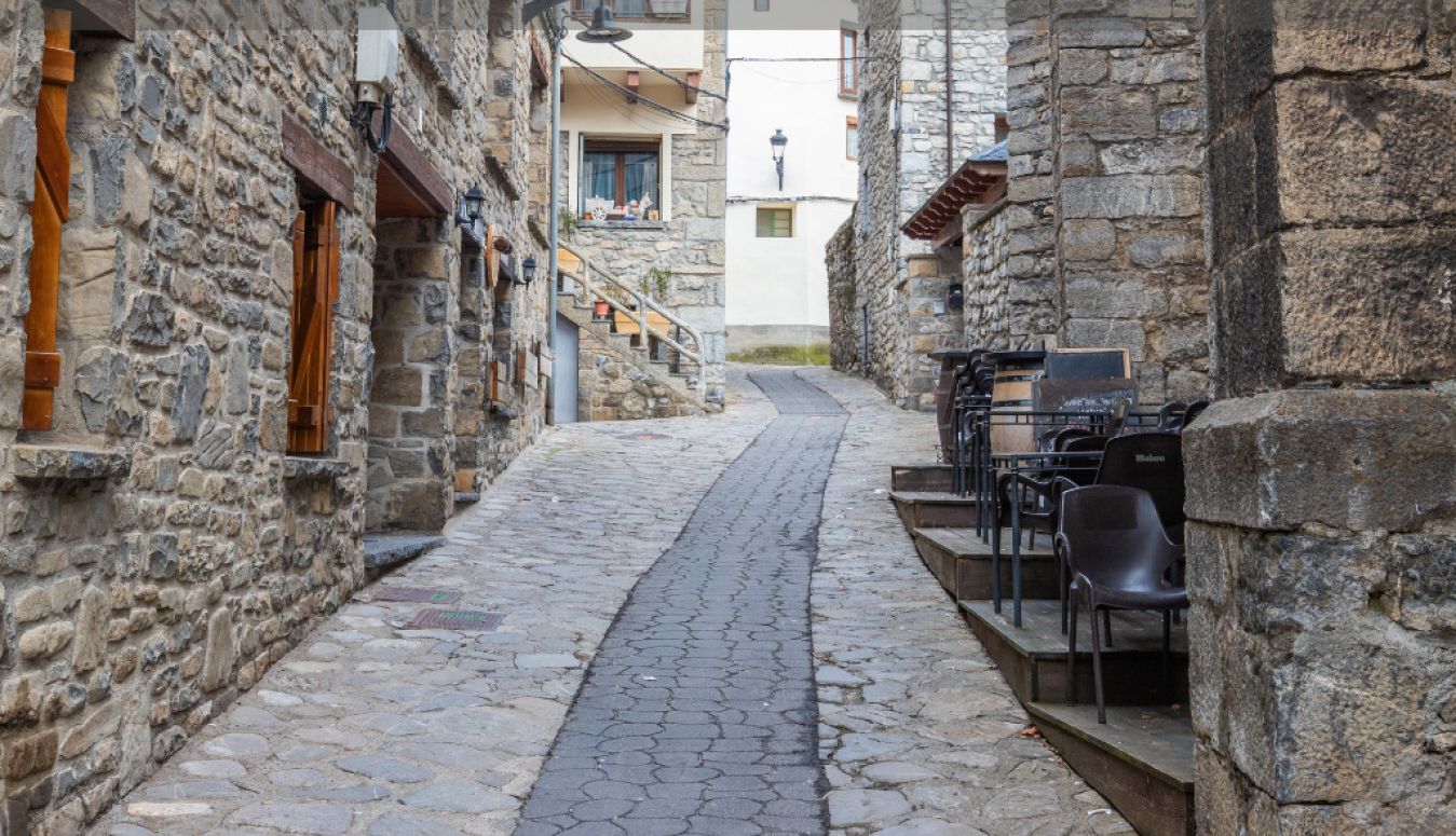 A narrow cobblestone street flanked by stone buildings with outdoor seating areas.