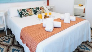 Un lit bien fait, agrémenté de serviettes, un plateau de jus d’orange et une table préparée pour le petit-déjeuner, le tout dans une pièce dont le sol est à carreaux et où le lit est recouvert d’une courtepointe blanche.