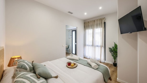 A hotel room with a bed, a TV mounted on the wall, a potted plant, and a window with curtains.