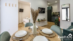 A modern living room with a dining table set for four, a sofa, a TV, and a kitchen area in the background.