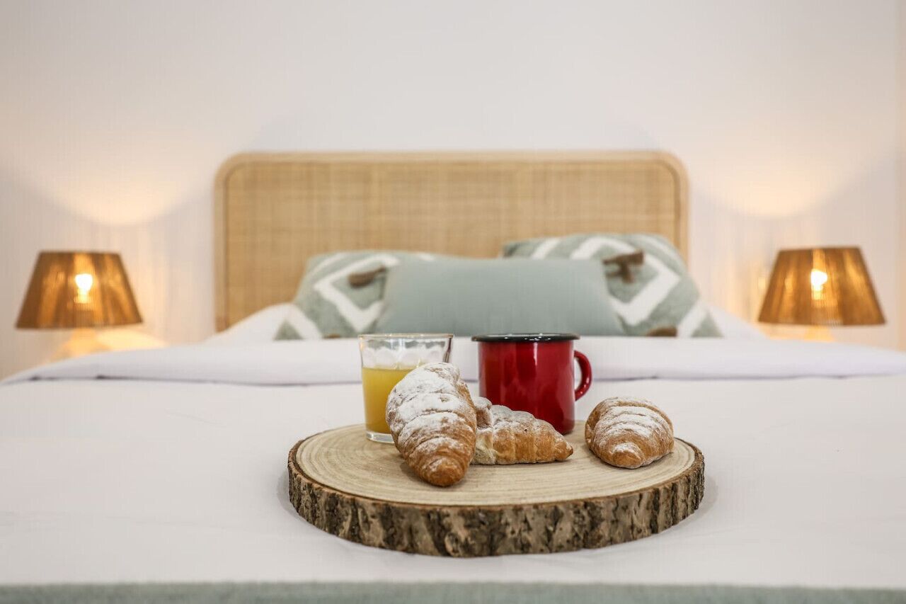 A bed with a wooden headboard, two lamps, a red mug, a glass of orange juice, and three croissants on a wooden coaster.