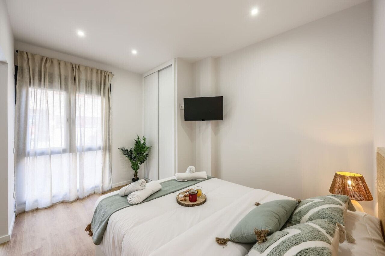 Un dormitorio con cama, televisor montado en la pared, ventana con cortinas y mesita de noche con lámpara.