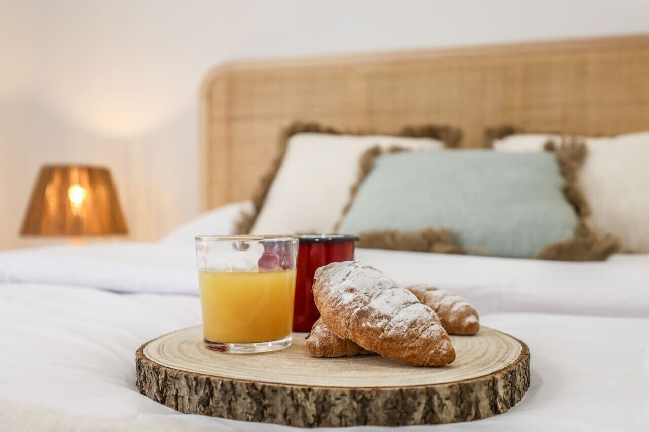 A breakfast setting with a glass of orange juice, a croissant, and a red mug on a wooden board on a bed with a headboard and pillows.