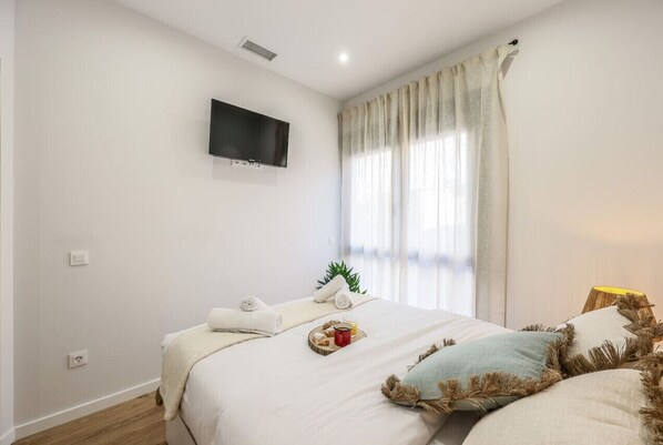 A bedroom with a bed, a flat-screen TV mounted on the wall, a window with curtains, and a bedside table with a lamp.