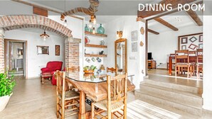 Een eetkamer met een bakstenen boog, houten stoelen, een glazen tafel en diverse decoratieve objecten op planken.