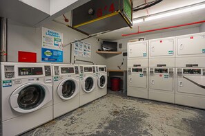 A laundry facility with multiple washing machines and dryers, a red trash bin, and a sign promoting easier laundry.