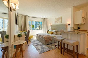 A modern hotel room with a bed, a desk, a chair, a small table with a plant, and a kitchen area with a microwave.