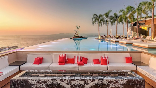 Zona junto a la piscina con sofá y sillones de descanso, vista al océano y palmeras.