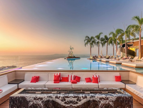 A poolside area with a sofa and lounge chairs, a view of the ocean, and palm trees.