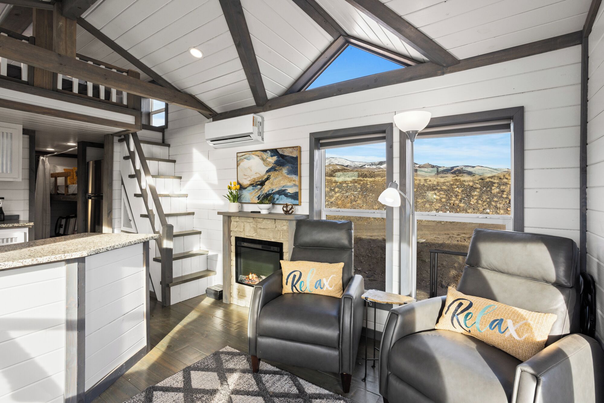 A modern living room with a fireplace, two leather chairs with 'Relax' pillows, a dining area, and a kitchen with a staircase.