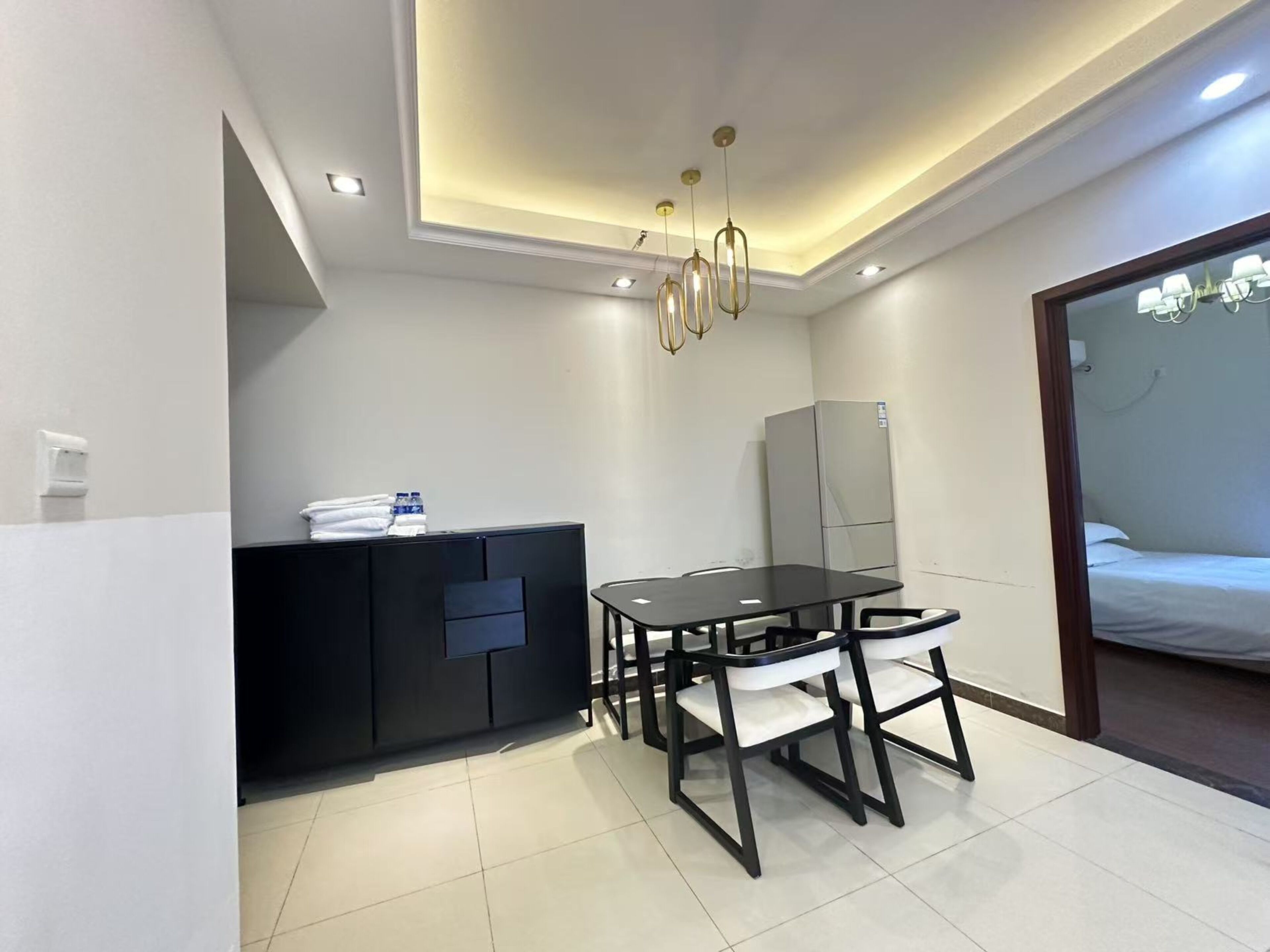 Una habitación de hotel moderna con comedor, un mueble negro con toalla y botella de agua, un refrigerador y una cama con cabecera blanca.