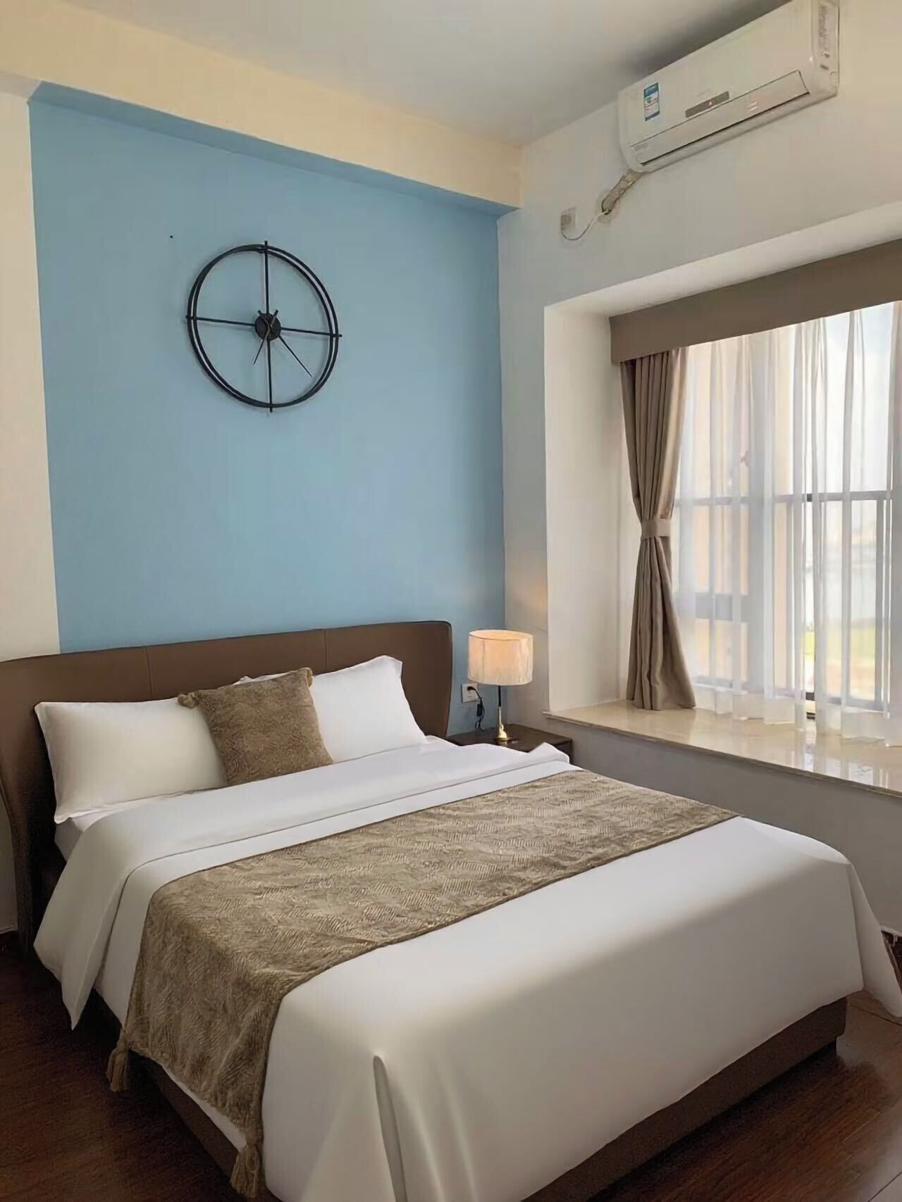 A hotel room with a bed, a wall clock, an air conditioner, a window with curtains, and a nightstand with a lamp.