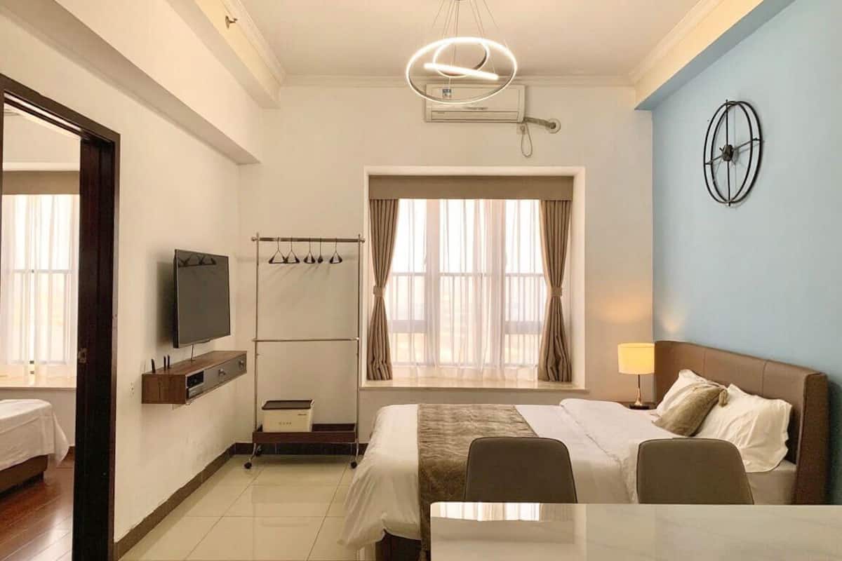 A hotel room with a bed, a TV, a window with curtains, a wall clock, and a ceiling light fixture.