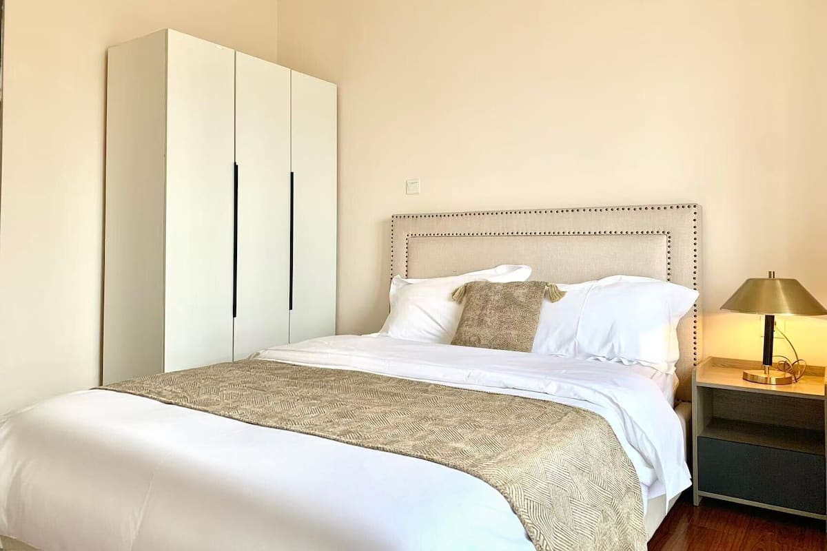 A neatly made bed with a patterned throw, a headboard with a decorative border, a bedside lamp, and a large wardrobe.