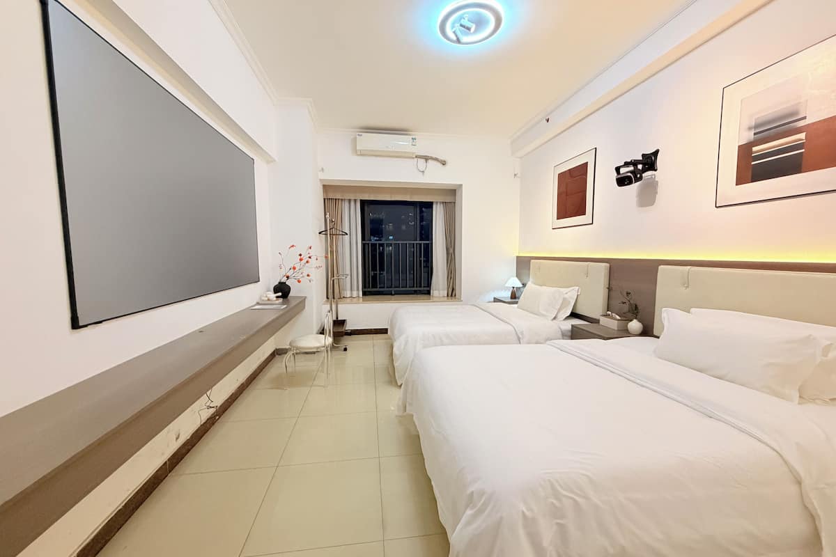 A hotel room with two beds, a flat-screen TV, a window with curtains, a bedside table with a lamp, and a ceiling light.