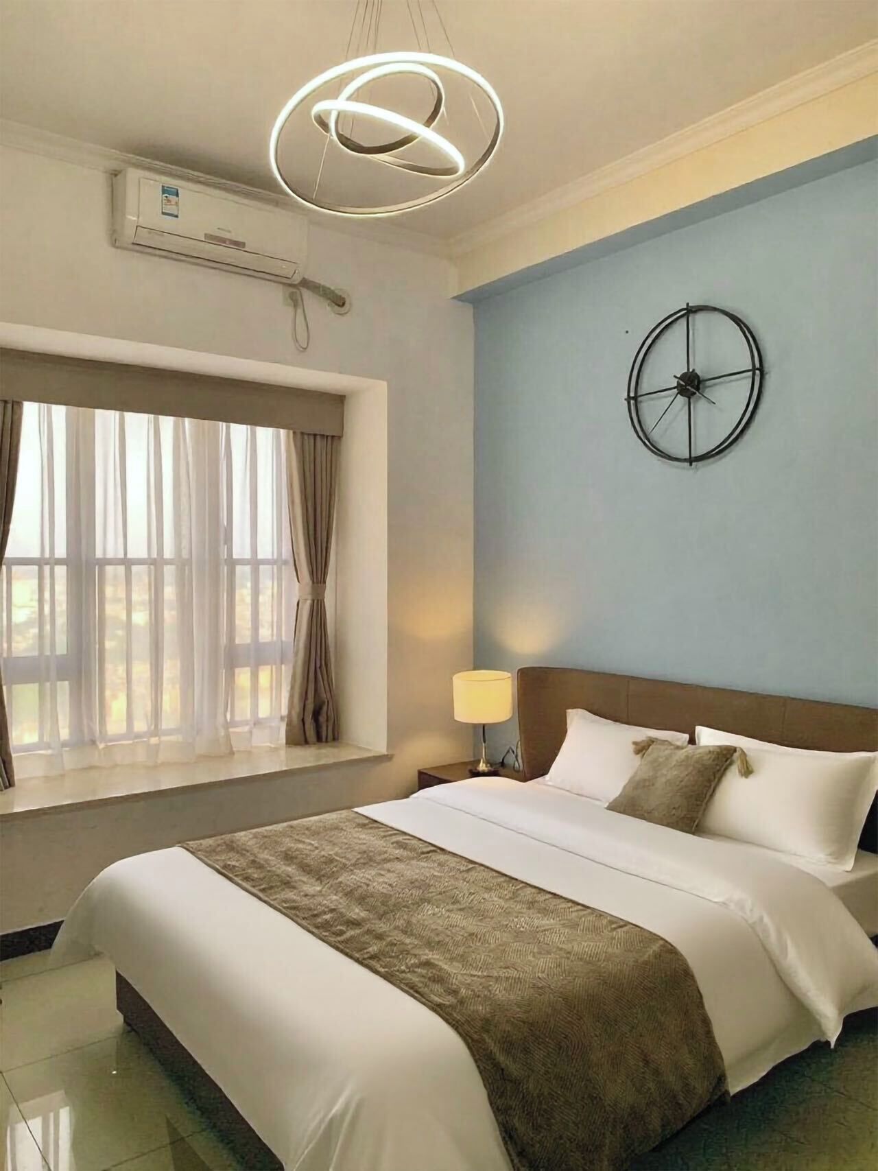 A hotel room with a bed, a window with curtains, a wall clock, and an air conditioning unit.
