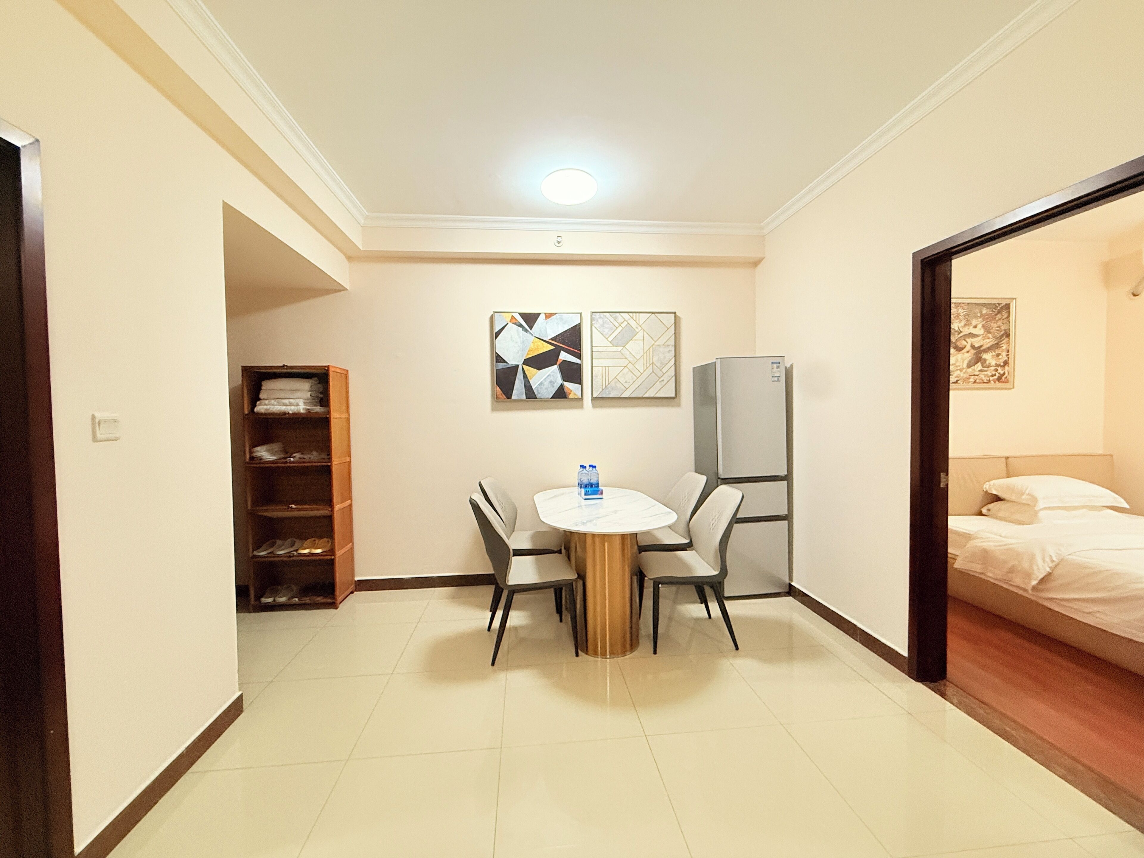 A compact hotel room with a bed, a small dining table with chairs, a refrigerator, and a bookshelf.