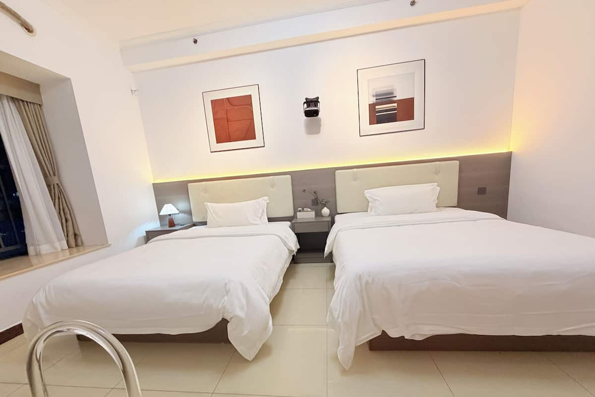 A hotel room with two beds, a nightstand with a lamp, a window with curtains, and two framed artworks on the wall.