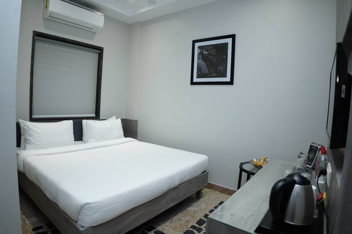 Ein Hotelzimmer mit einem Bett, einem kleinen Tisch mit Wasserkocher, einem gerahmten Bild an der Wand und einer Klimaanlage über dem Bett.