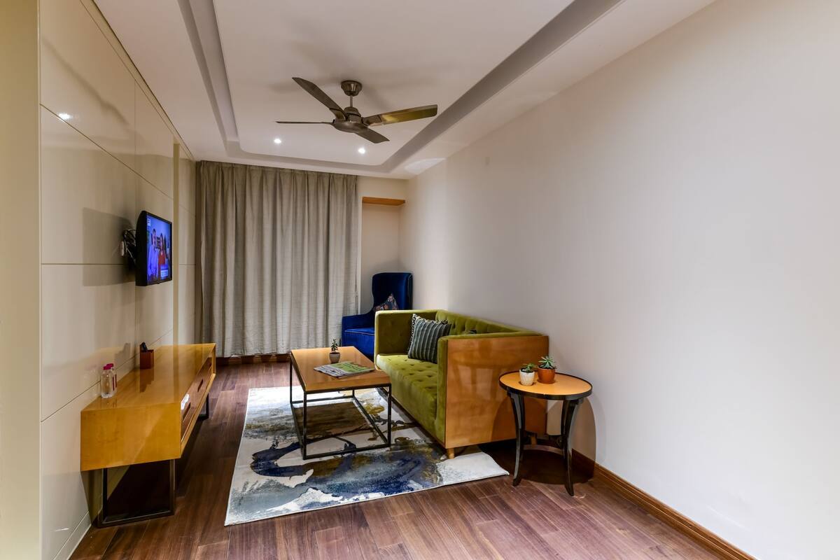 Ein modernes Wohnzimmer mit einem grünen Sofa, einem Holztisch, einem kleinen runden Tisch, einem an der Wand befestigten Fernseher und einem Deckenventilator.