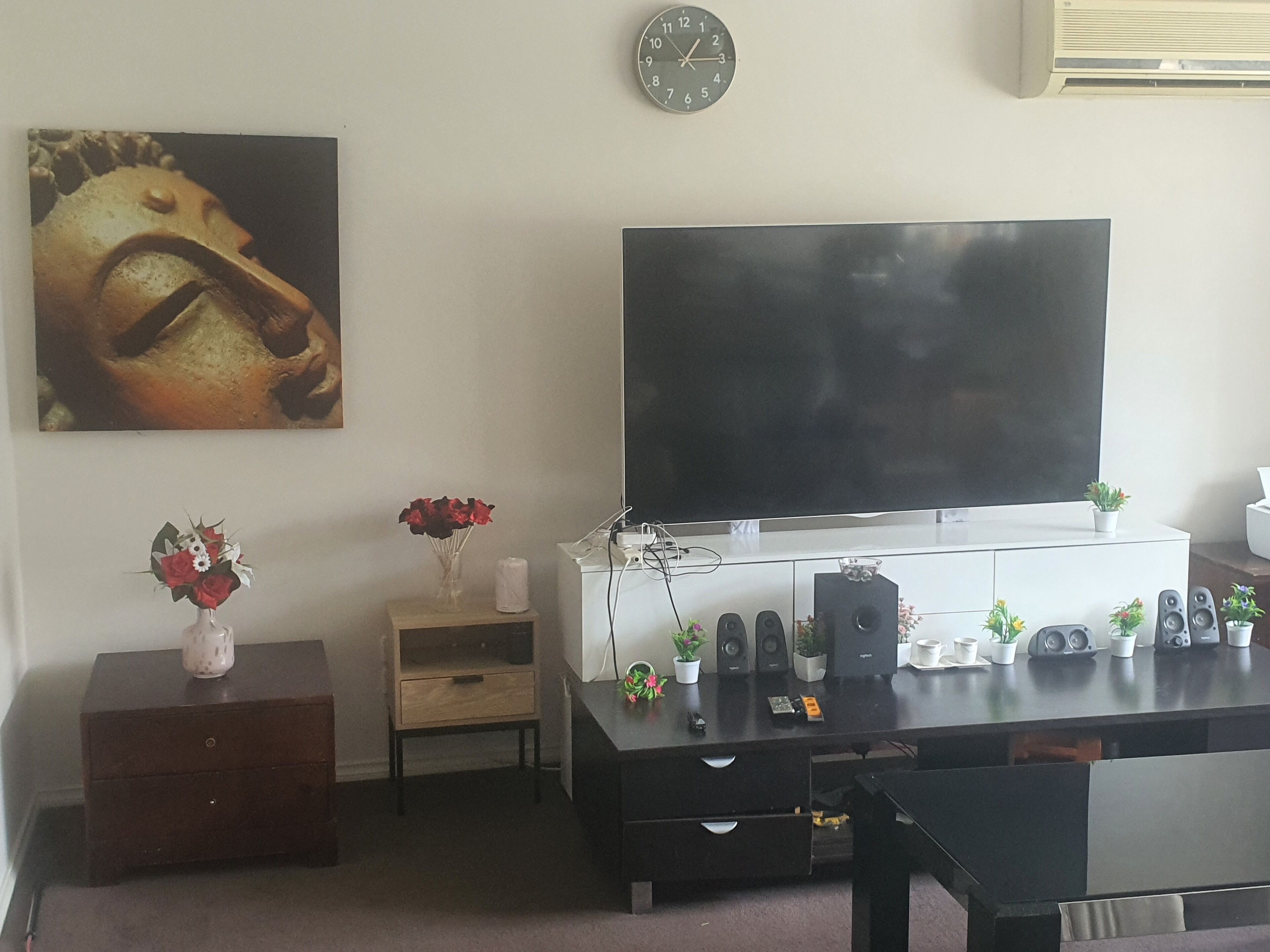A living room with a large flat-screen TV mounted on the wall, a wooden cabinet, a small side table with a vase of flowers, and a clock on the wall.