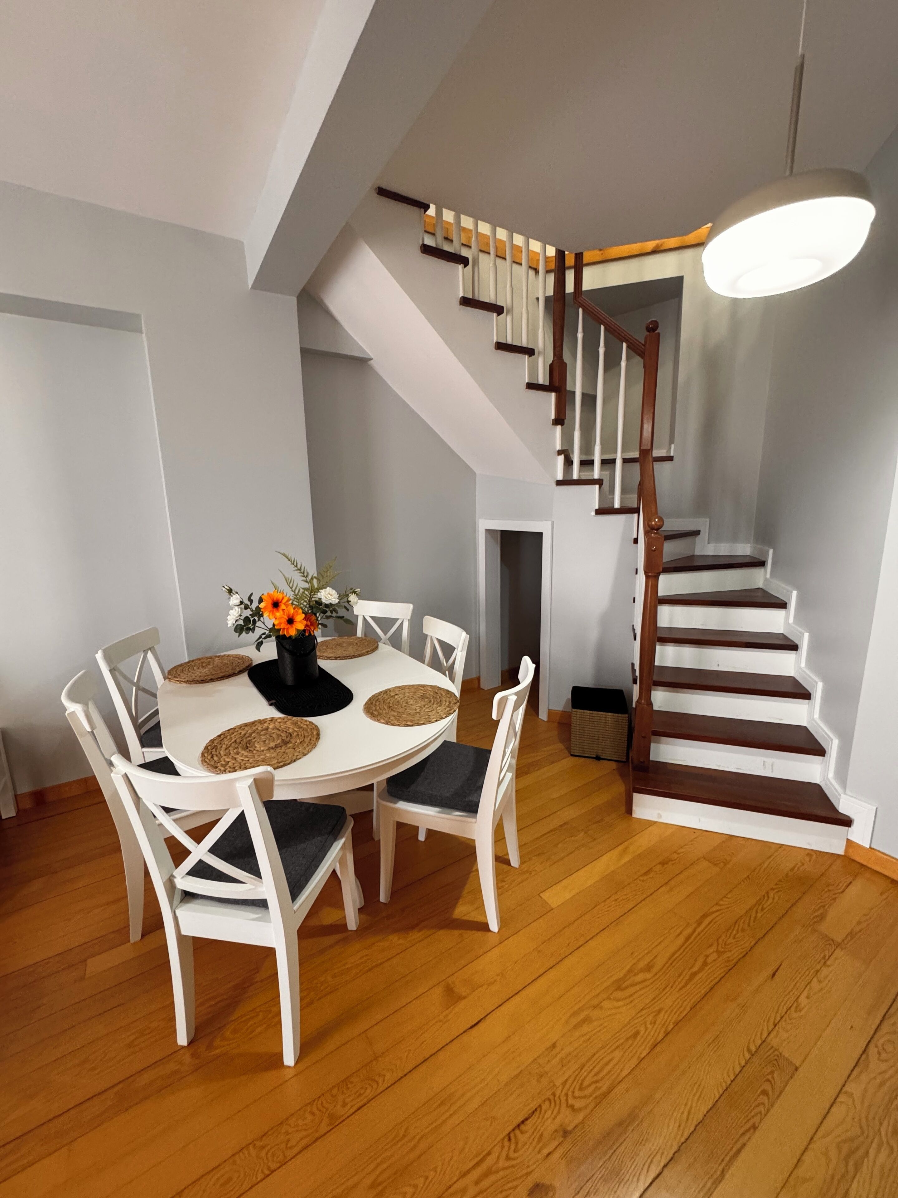 A dining area with a round table, white chairs, and a vase with flowers. A wooden staircase with a railing leads to an upper level.