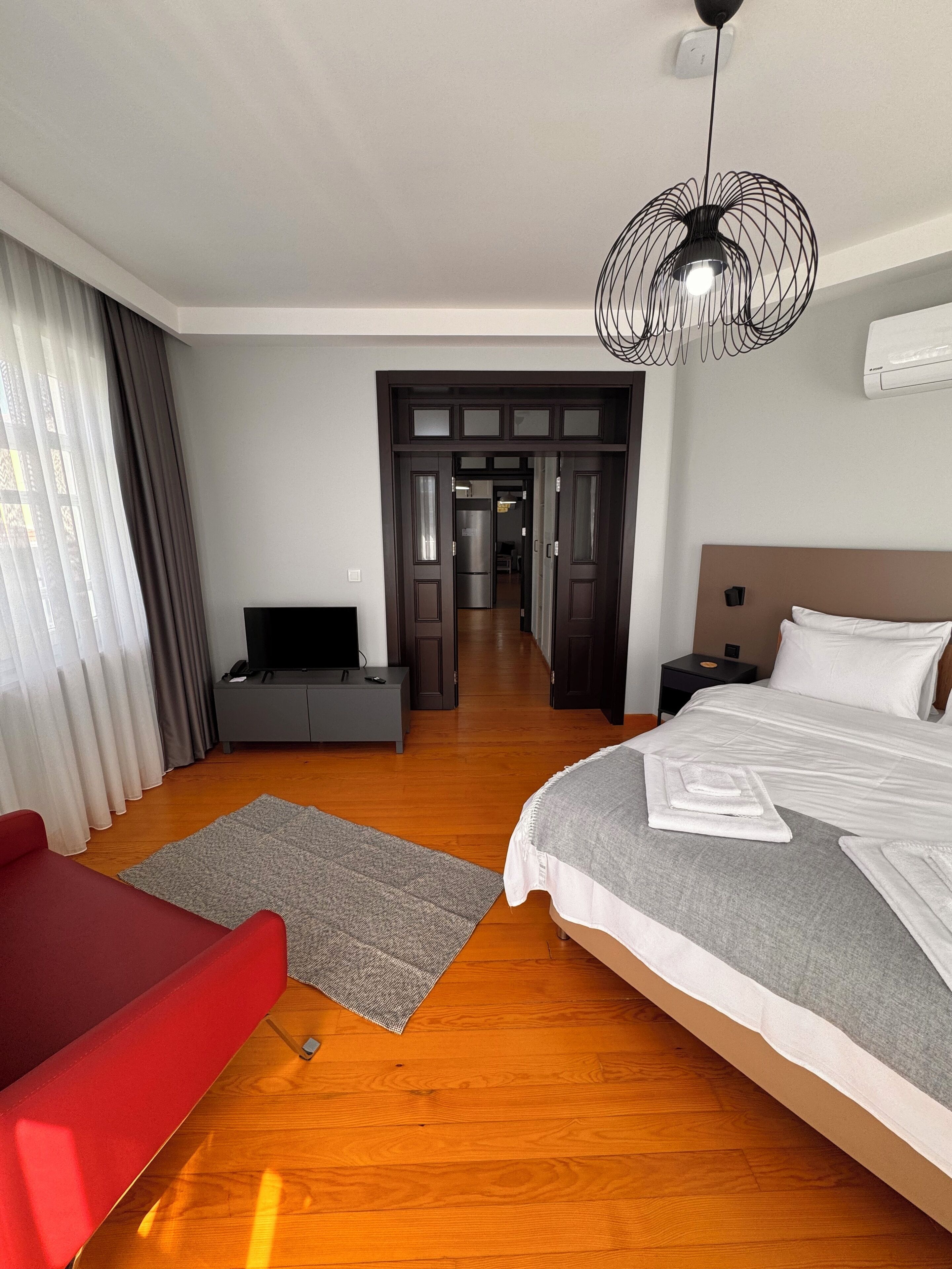 A hotel room with a large bed, a red armchair, a grey sofa, a TV, and a modern pendant light.