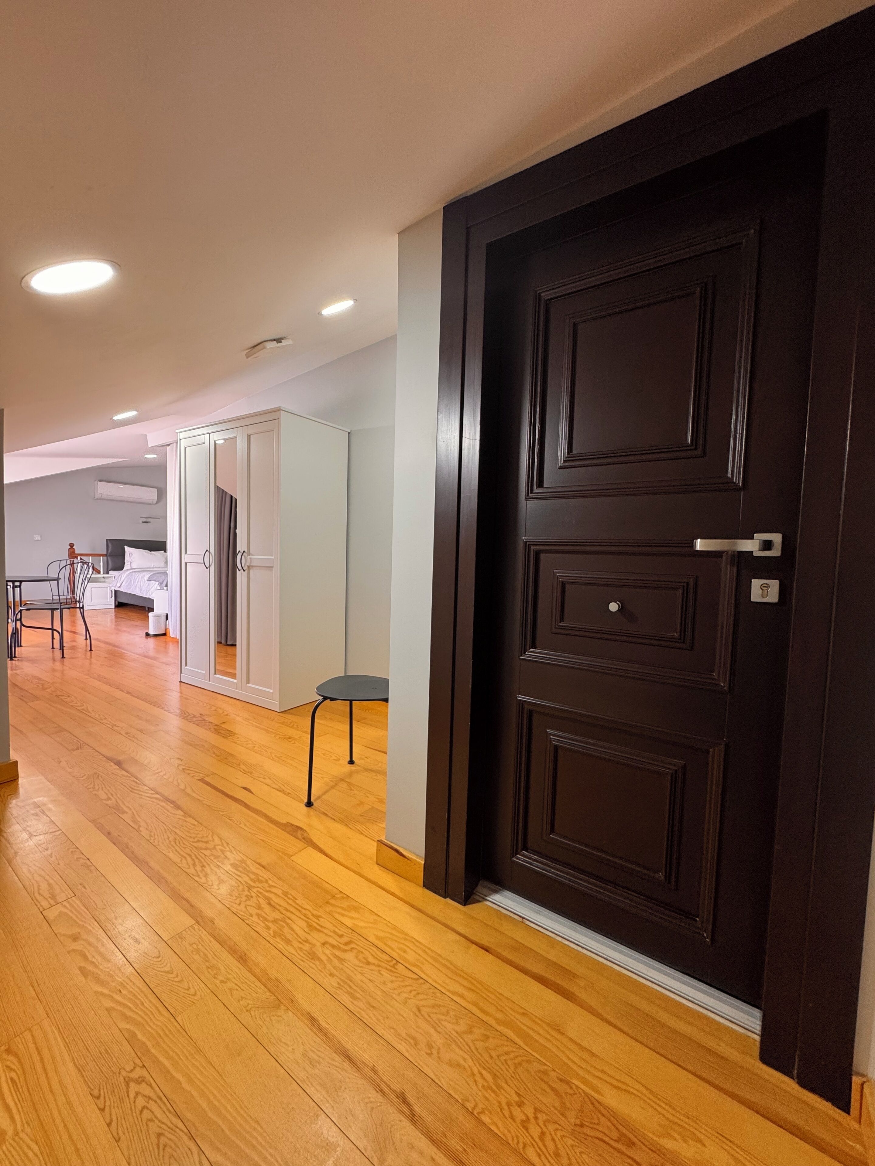 A hotel room with a wooden floor, a dark wooden door, a bed, a desk, and a chair.