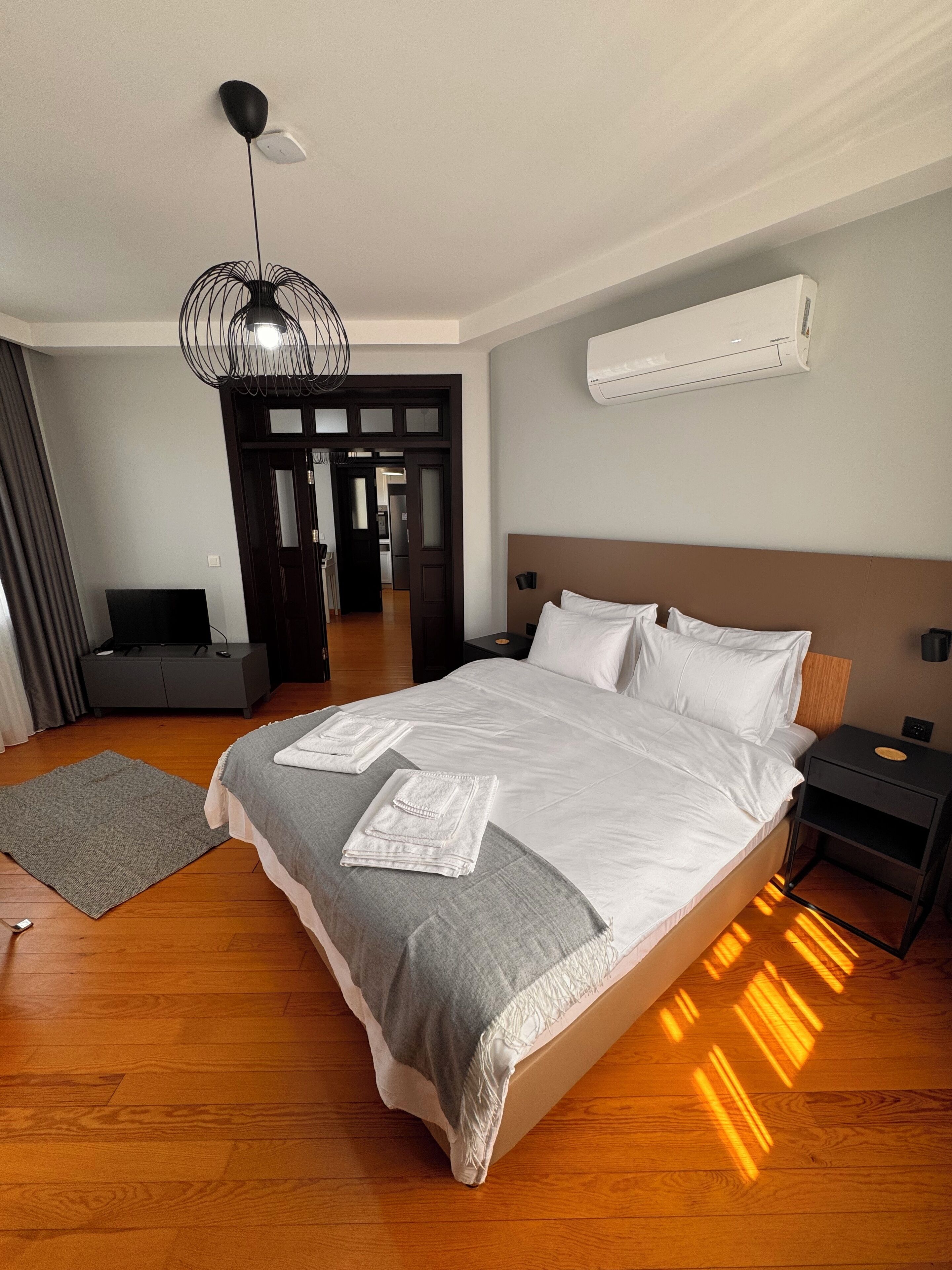Moderni hotellihuone, jossa on suuri sänky, harmaa heittopeitto, harmaa matto, musta yöpöytä, musta lamppu ja valkoinen ilmastointilaite.