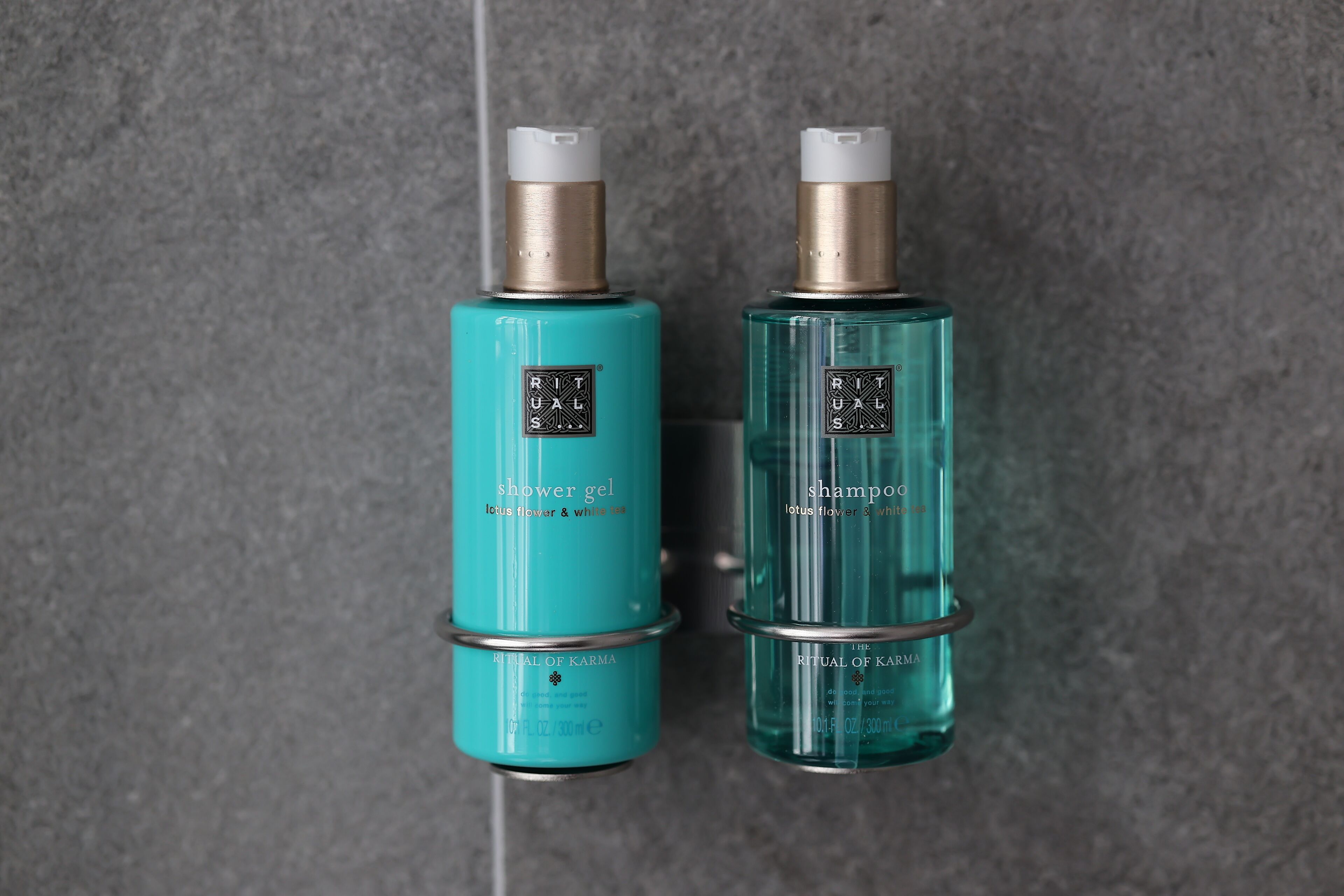 Two bottles of Ritual of Karma products, one labeled 'shower gel' and the other 'shampoo', both in teal and clear glass with metallic caps.