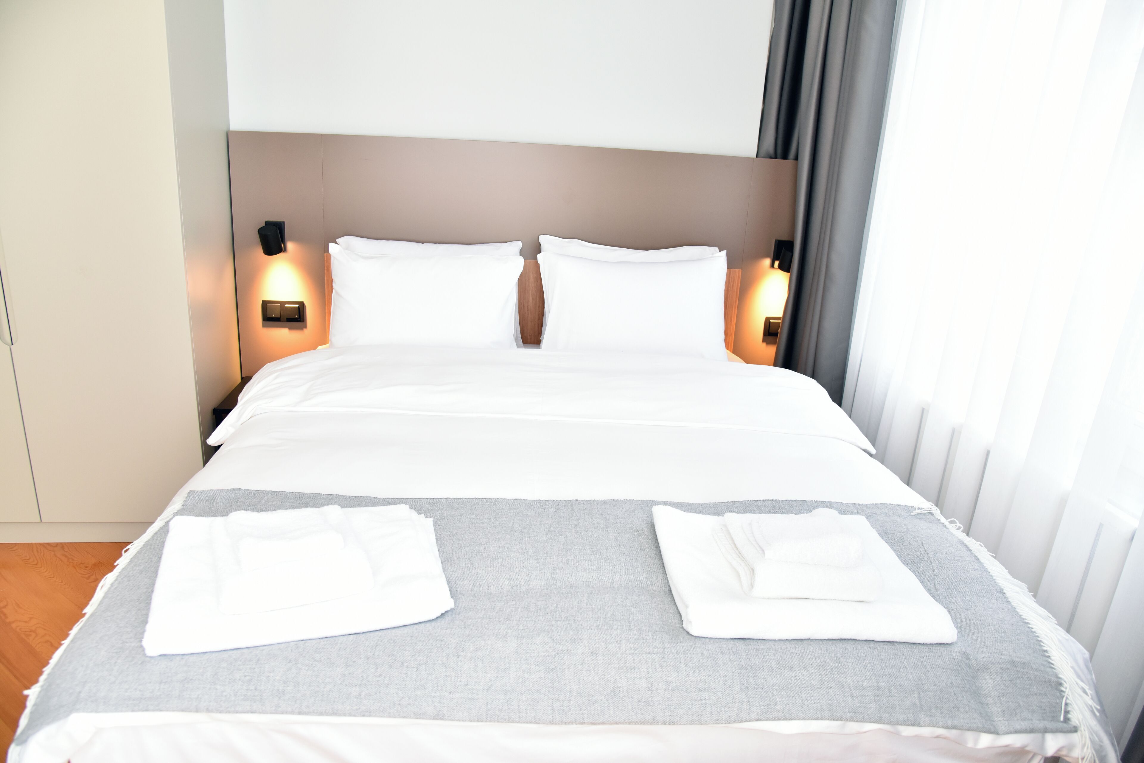 A neatly made bed with white linens, two pillows, and a grey blanket. There are two bedside lamps on either side of the bed.